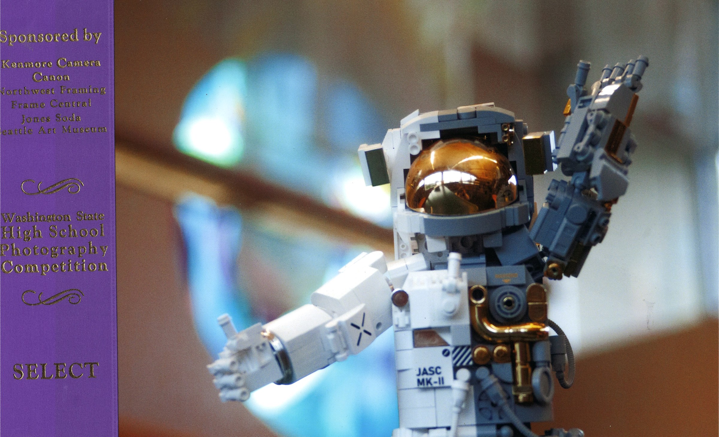 Photograph of a Lego astronaut figure posed against a blue background, mounted with a purple art show ribbon