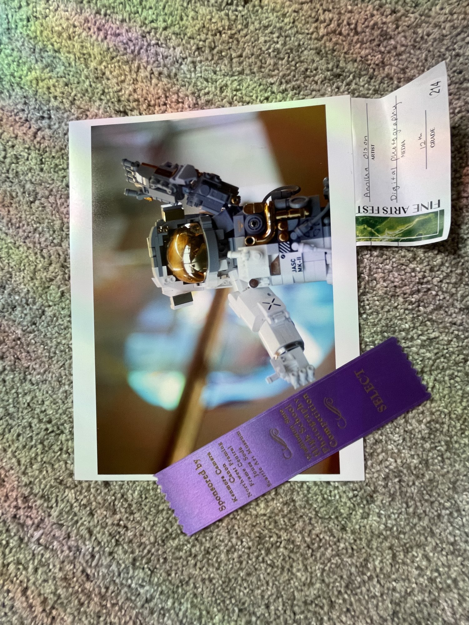 Photograph of a Lego astronaut figure posed against a blue background, printed and mounted with a purple fair ribbon